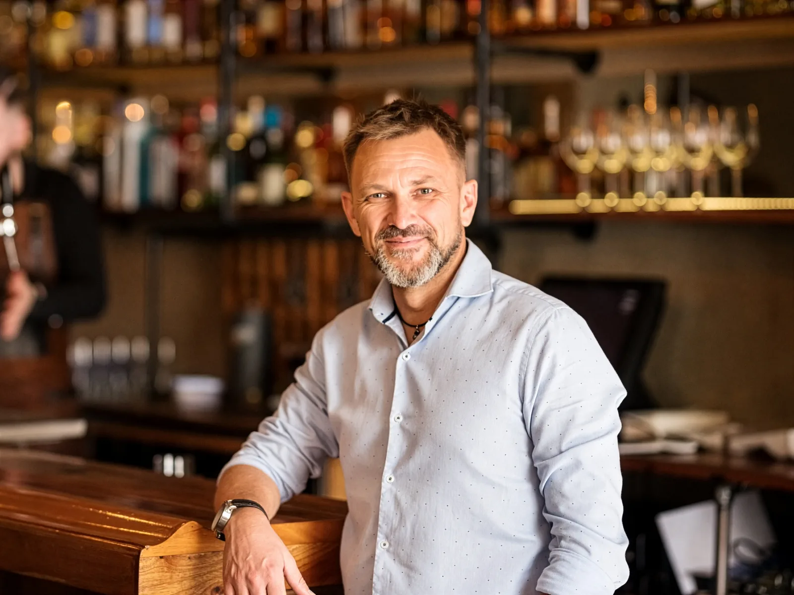 A bar owner leaning on his counter, with the bartender working behind him.